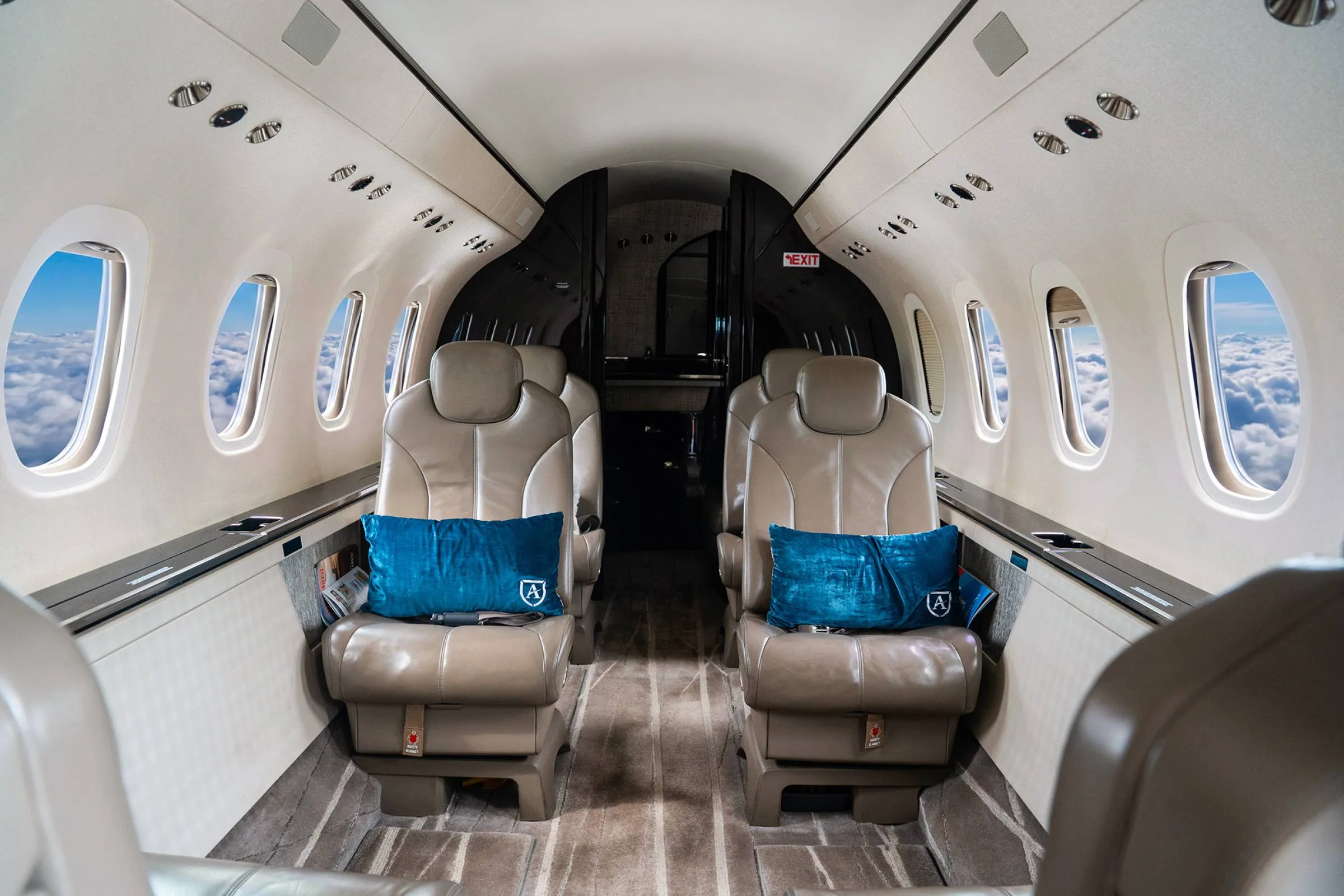The luxurious interior cabin of a private jet features tan leather seats with blue pillows, an "EXIT" sign, and windows showing clouds.