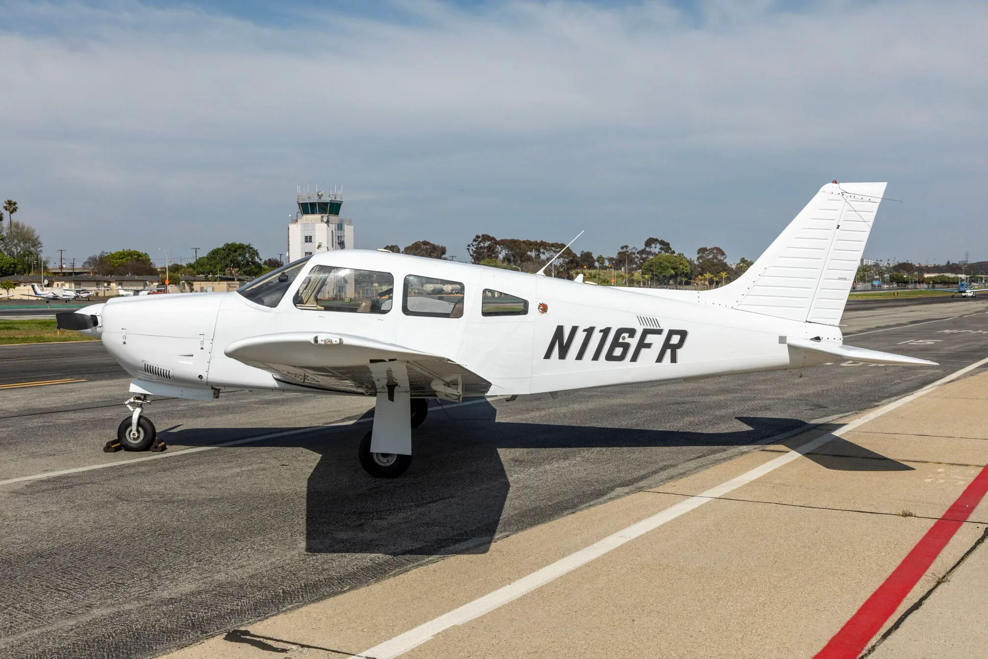 A white Piper PA-28-181 Archer III aircraft, registration N116FR, parked on an airfield with a control tower in the background.