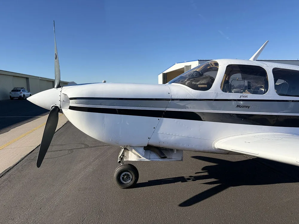 A close-up front-left view of the propeller and nose of a white Mooney aircraft with black and grey stripes.