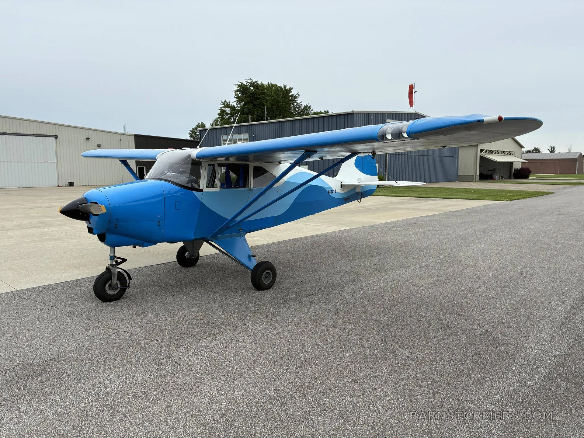 A blue and light blue Piper Cub-style aircraft with a wavy paint scheme is parked on an asphalt ramp in front of hangars under a cloudy sky.