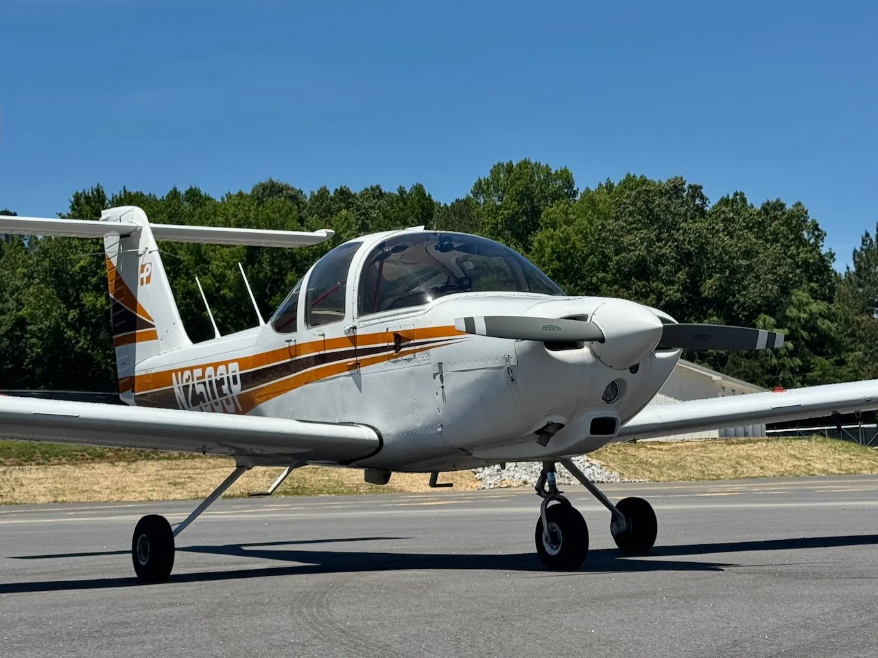 A white Piper PA-38 Tomahawk aircraft with orange and black stripes, seen from the front-right on an airfield.