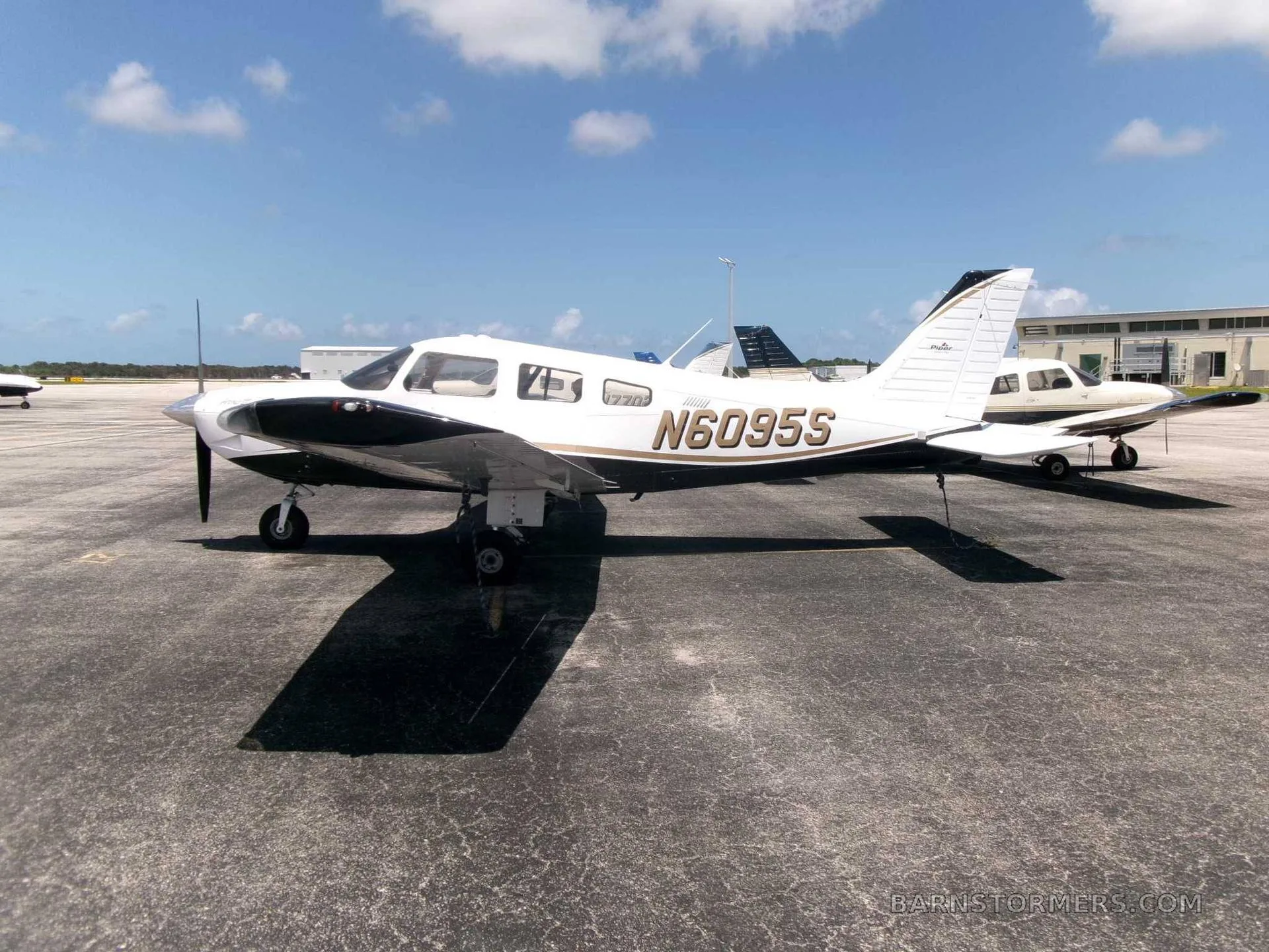 A white and black Piper PA-28 Cherokee with gold trim, registration N6095S, parked on an asphalt ramp under a partly cloudy sky.