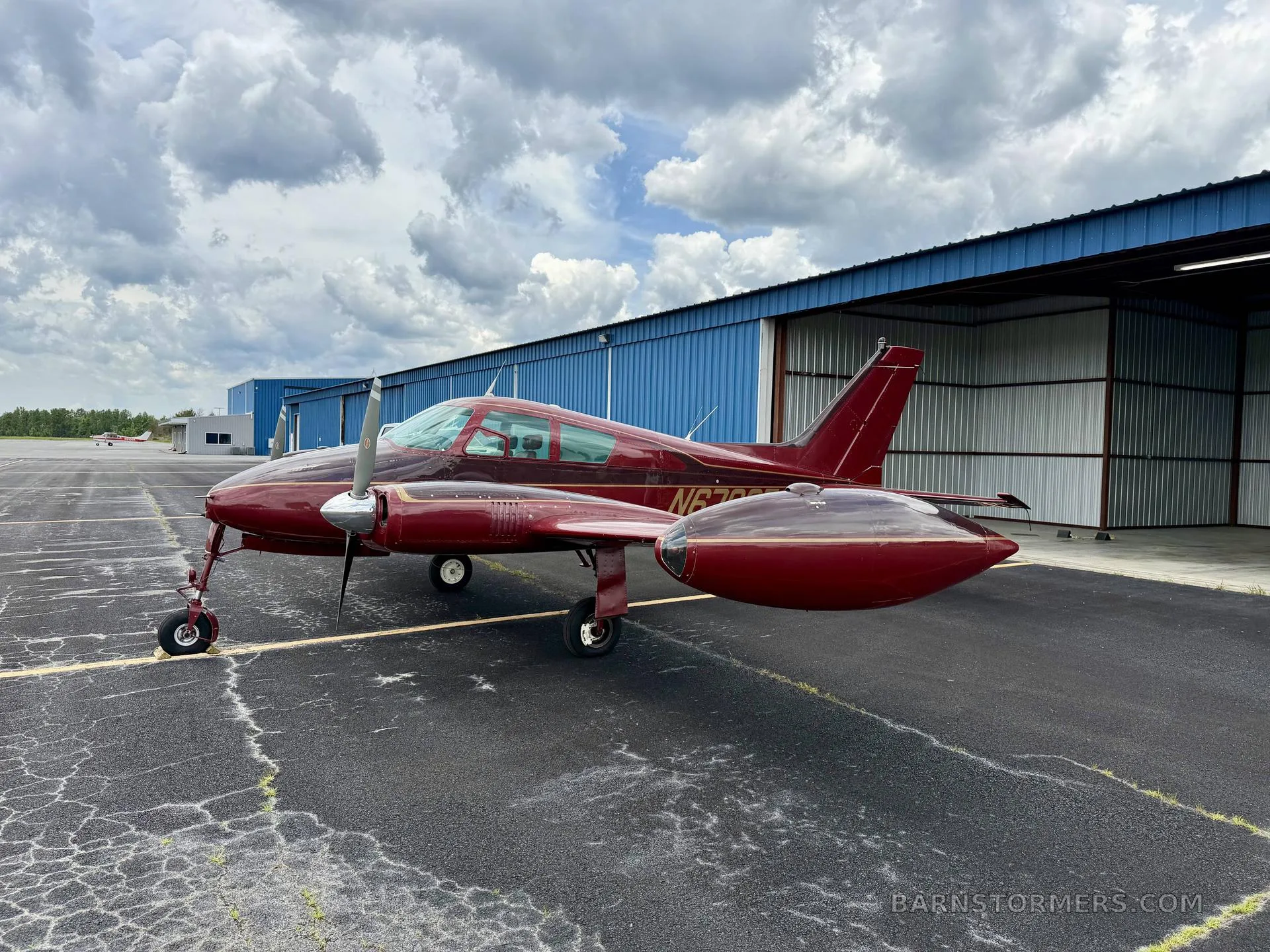 A red twin-engine aircraft, likely a Beechcraft Baron, with registration N6792T, parked on an asphalt apron next to a blue hangar under a cloudy sky.