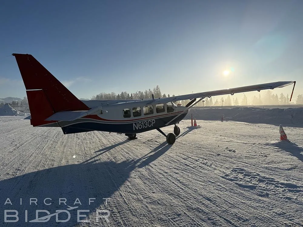 A rear-side view of the N613CP Cessna 206, blue and white with a red tail, parked on a snowy field with the sun setting in the background.