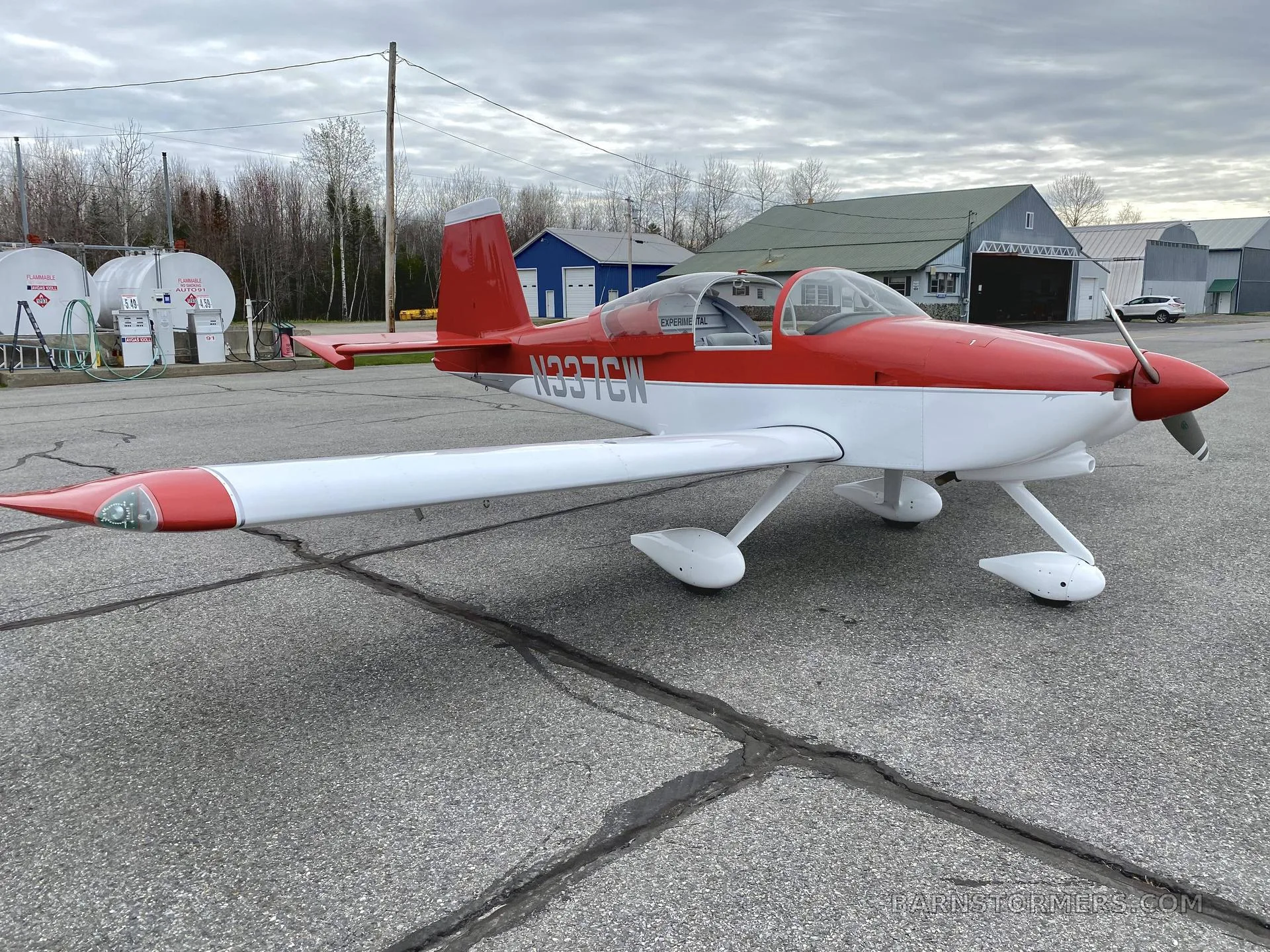 A red and white experimental aircraft with registration N337CW is parked on an airfield with fuel tanks and buildings in the background.