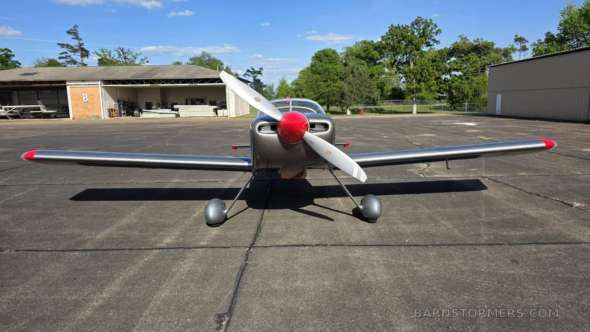 A front view of a silver single-engine aircraft with a red propeller spinner and red wingtips, parked on an asphalt tarmac with trees and a building in the background.