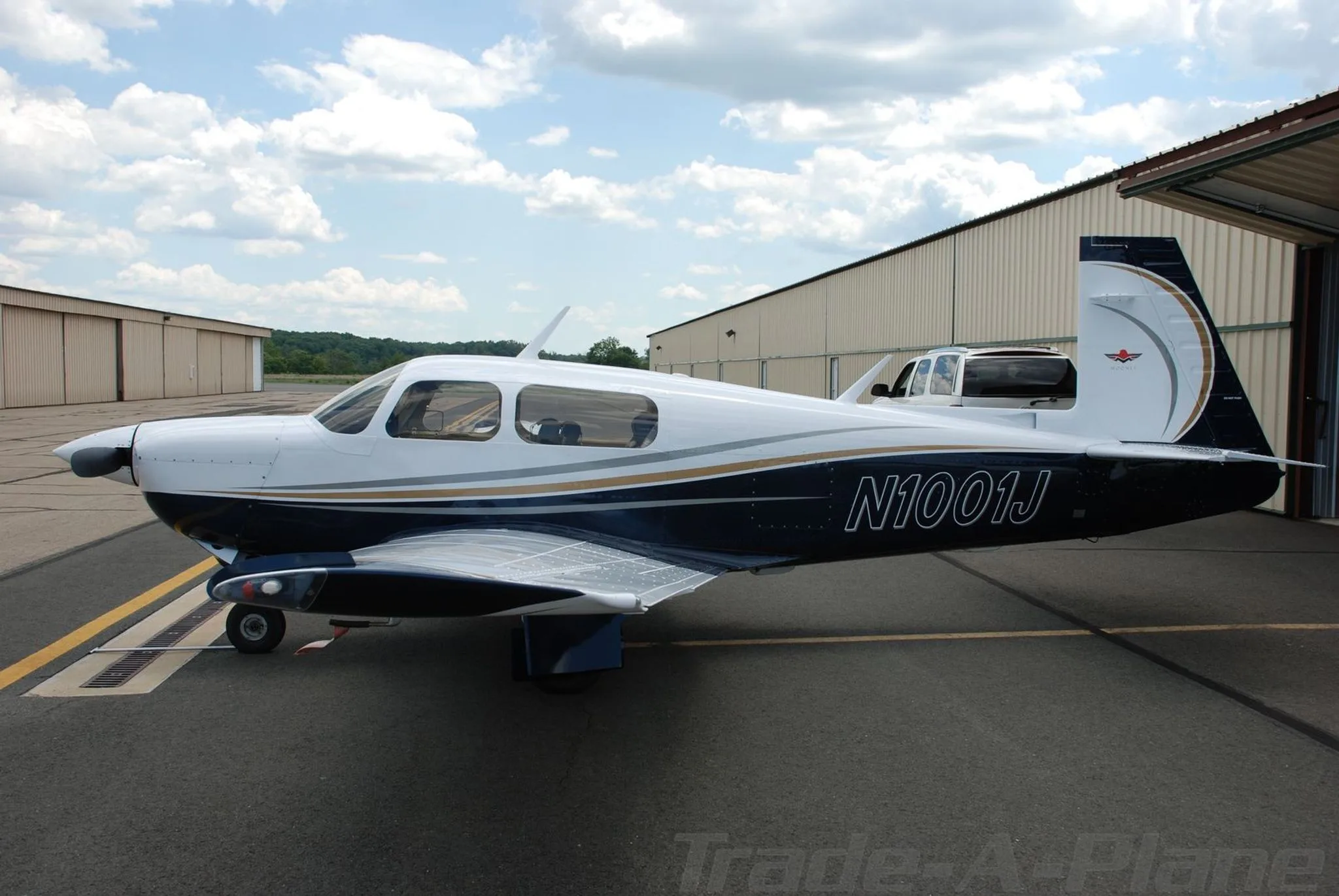 A side view of the white and blue Mooney aircraft (N1001J) parked on a tarmac next to a hangar.
