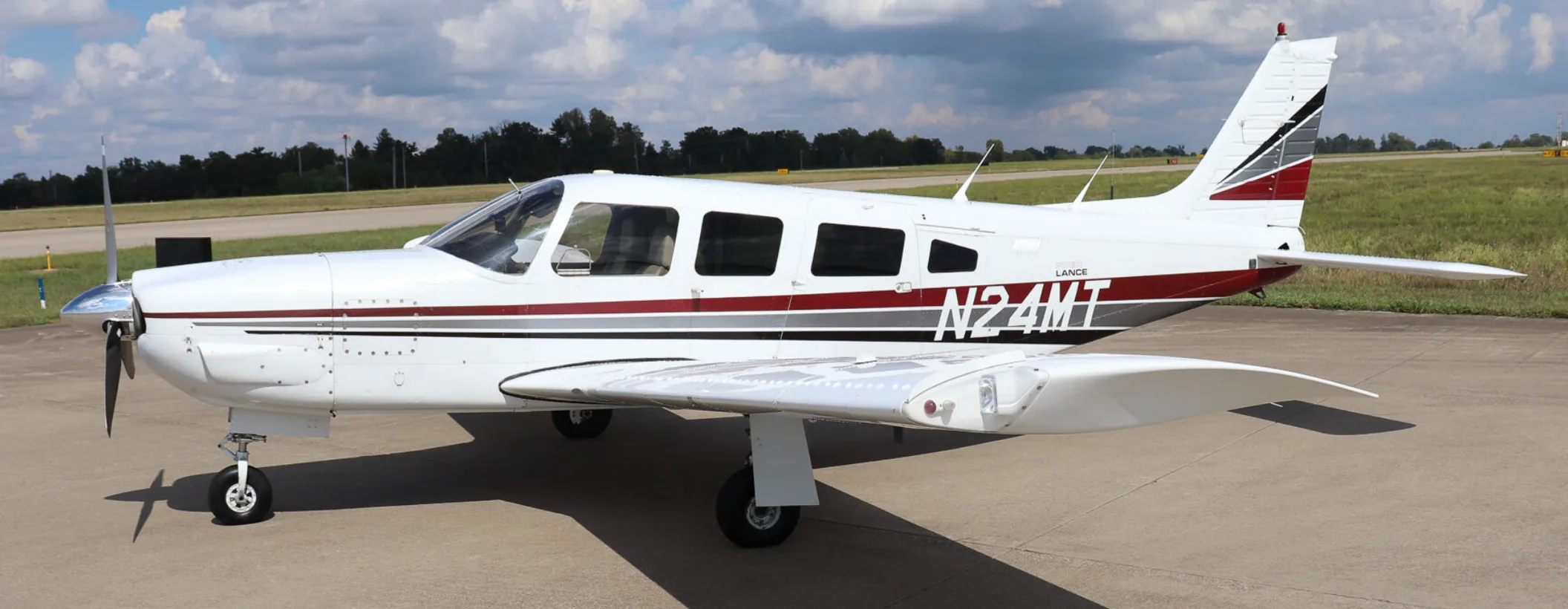 A white Piper Lance (N24MT) with red and grey stripes is parked on an airfield, viewed from the left side.