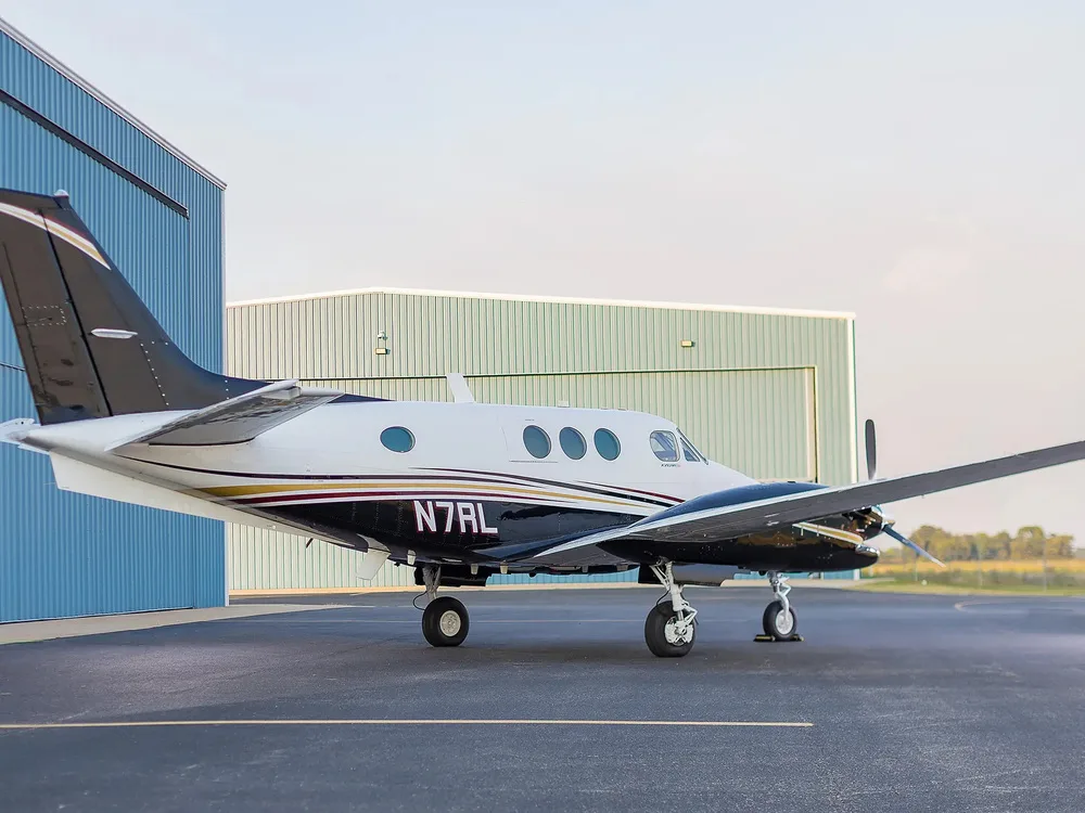 A side view of the N7RL Beechcraft King Air aircraft, white and black with gold and red stripes, parked on an tarmac.