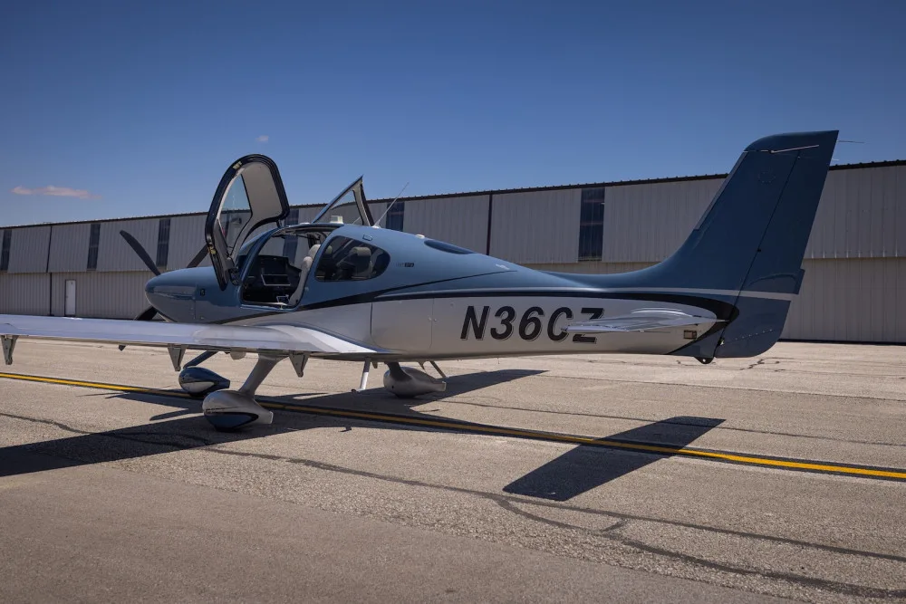A blue and silver Cirrus SR22T aircraft with open doors parked on an airport tarmac, viewed from the rear-side.