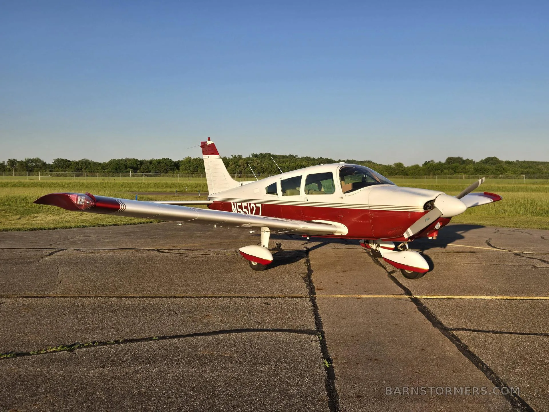 A red and white single-engine propeller aircraft (N55127) is parked on an asphalt ramp, viewed from the front-right side under a clear blue sky.
