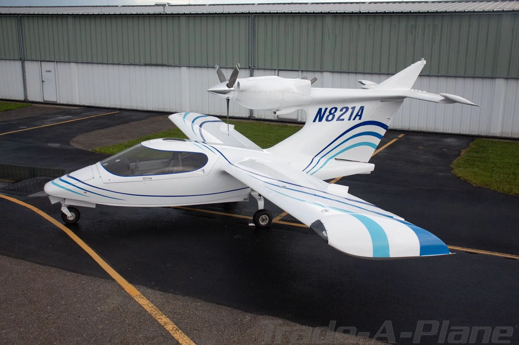A white Searey amphibian aircraft with blue stripes, registration N821A, parked on a wet tarmac next to a hangar.