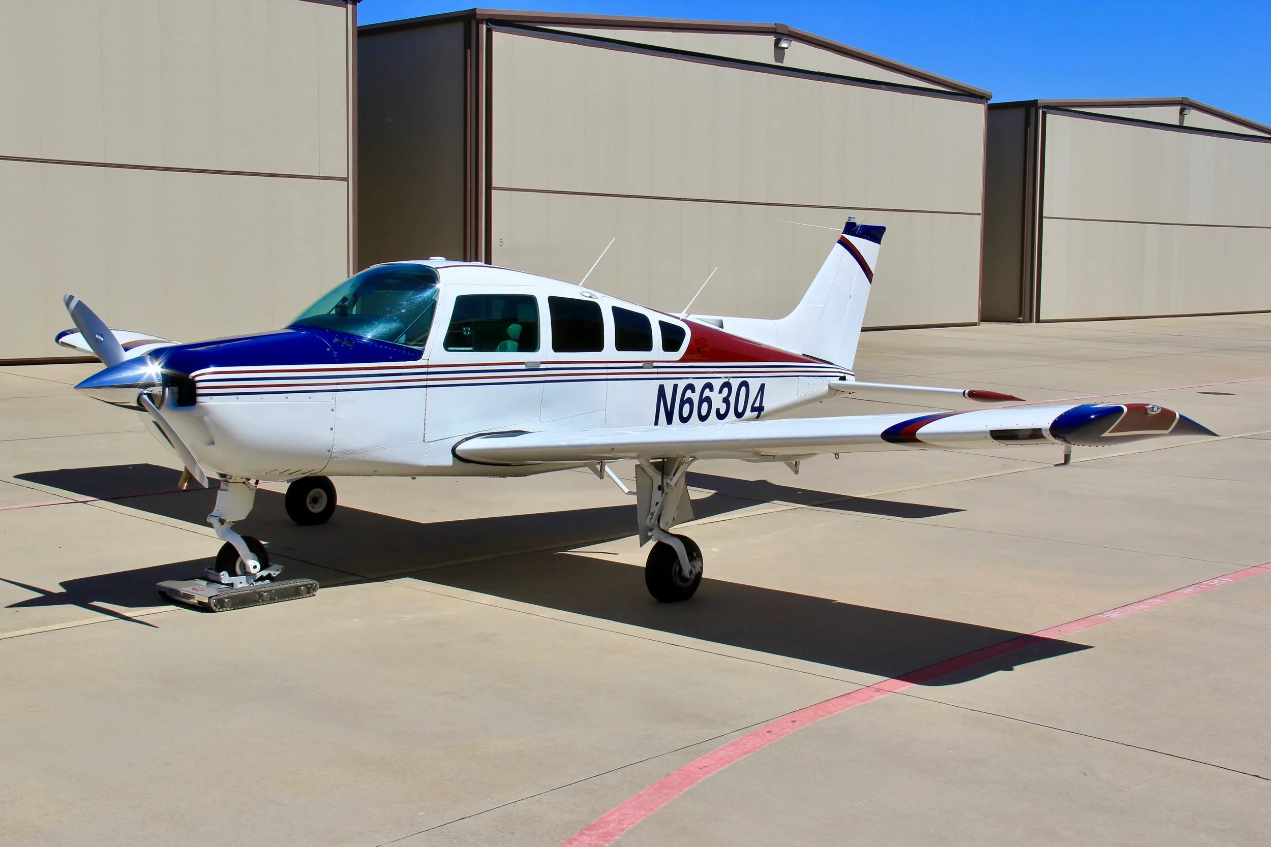 A white, blue, and red single-engine aircraft (N66304) parked on an tarmac in front of hangars.