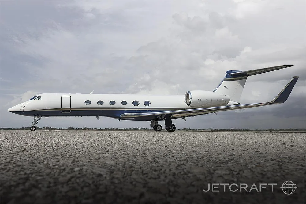 A white and blue Gulfstream G550 business jet is parked on a tarmac under a cloudy sky.