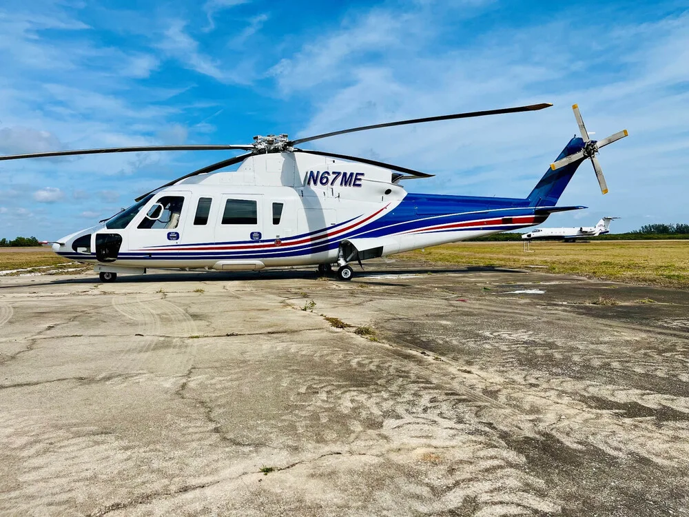 Side view of a white and blue helicopter (N67ME) with red stripes parked on a concrete pad.