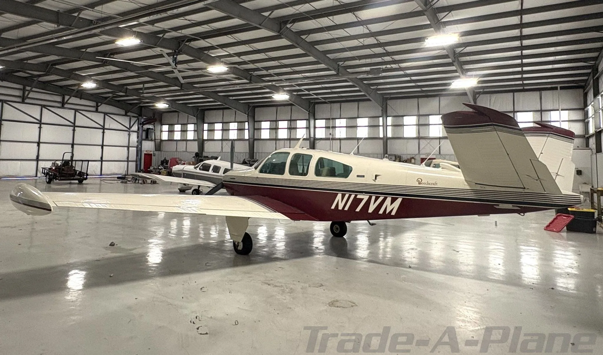 A white and maroon Beechcraft Bonanza N17VM parked inside a hangar, viewed from the rear-right.