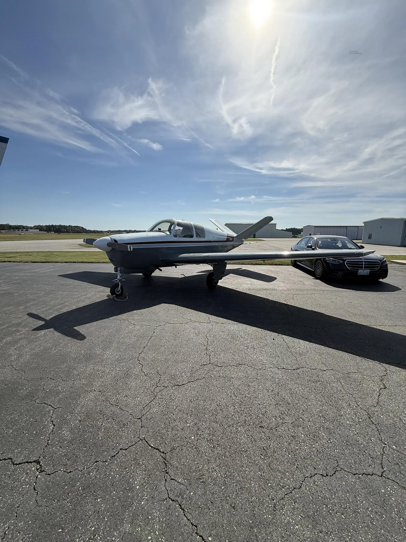 The N2194D Beechcraft Bonanza V-tail aircraft, with a white and grey paint scheme and red stripe, parked on an airfield tarmac next to a black car.