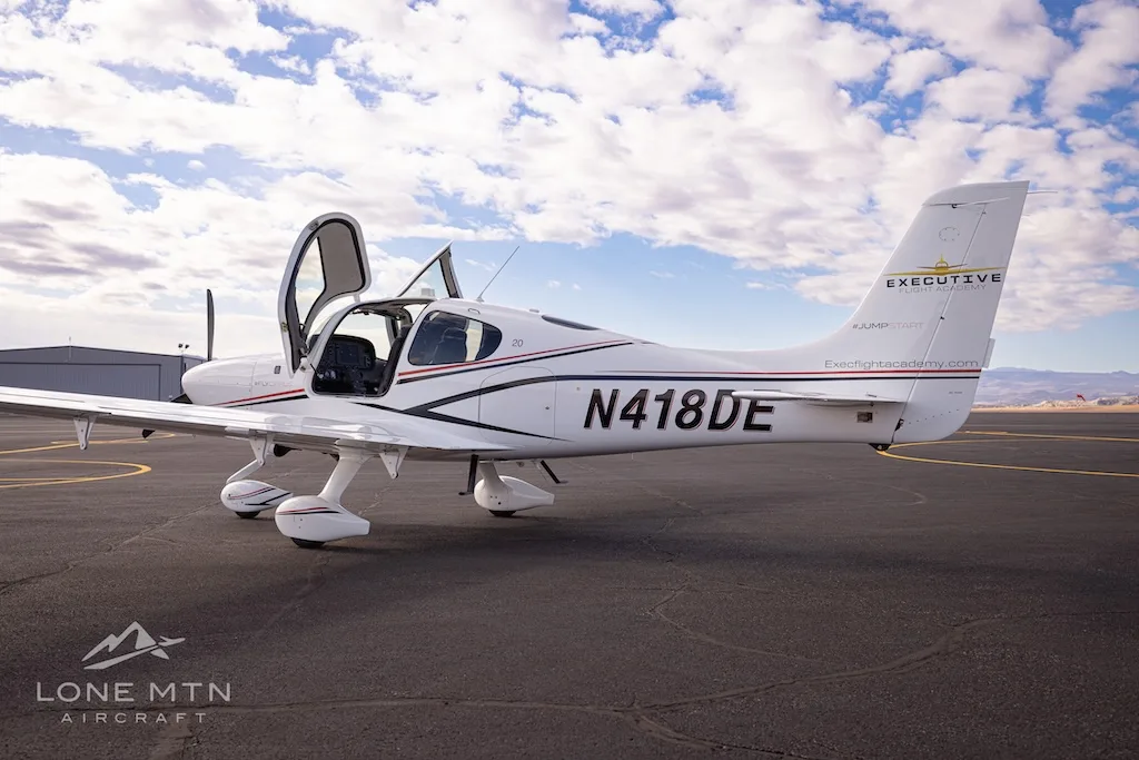 A white Cirrus SR22 aircraft with black and red stripes, registration N418DE, parked on an airfield with doors open.