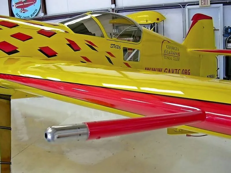 A close-up view of the yellow and red Sukhoi Su-26 aircraft's fuselage and wing, featuring a red pitot tube, inside a hangar.