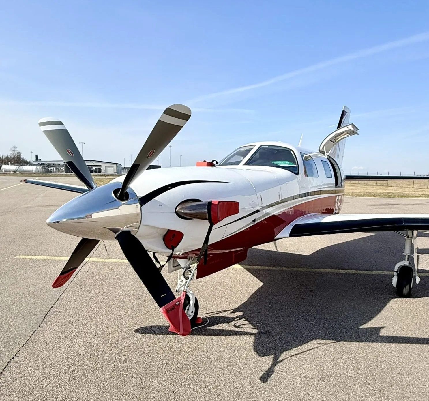 A white and red single-engine turboprop aircraft with its passenger door open, parked on an airport tarmac under a clear blue sky.