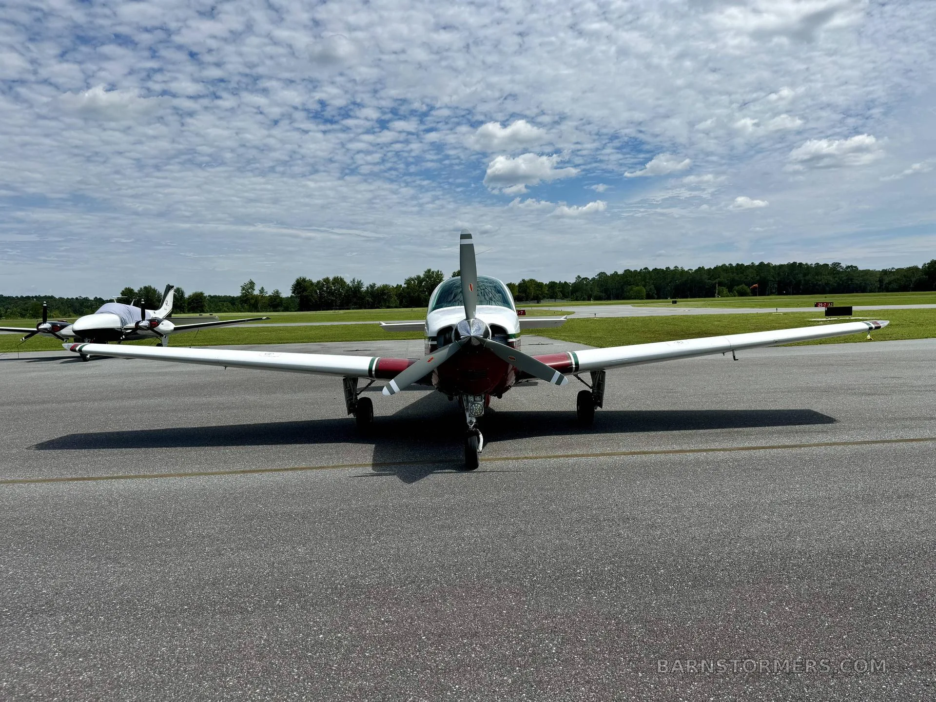 A white and maroon single-engine propeller aircraft is viewed head-on on an airfield under a cloudy sky, with another aircraft in the distance.