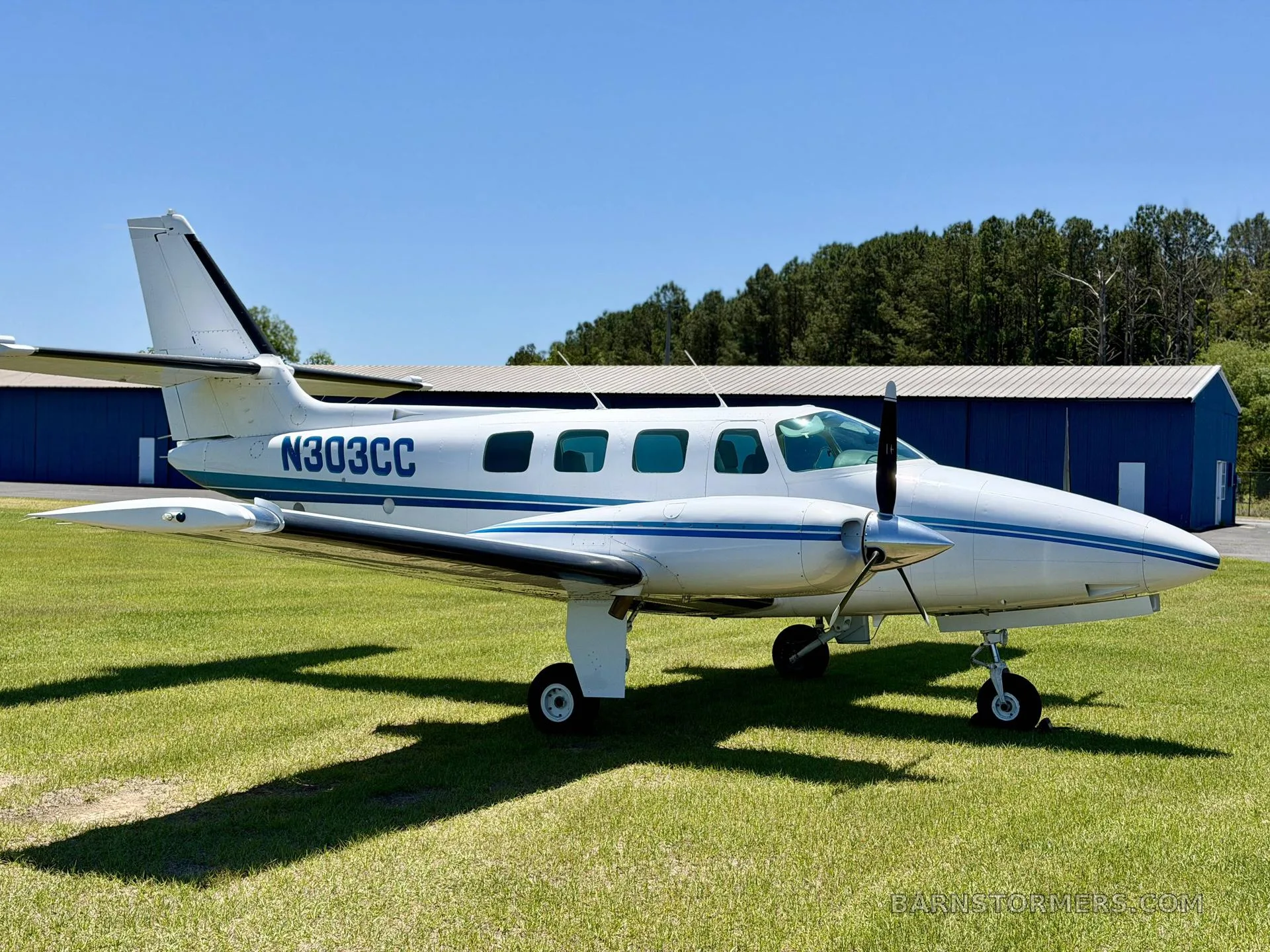 A white twin-engine propeller aircraft with blue stripes (N303CC) is parked on a grassy field, facing a blue hangar.