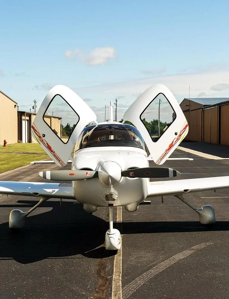 A front view of a white Cirrus SR20 aircraft with open doors and a two-blade propeller on an airfield.