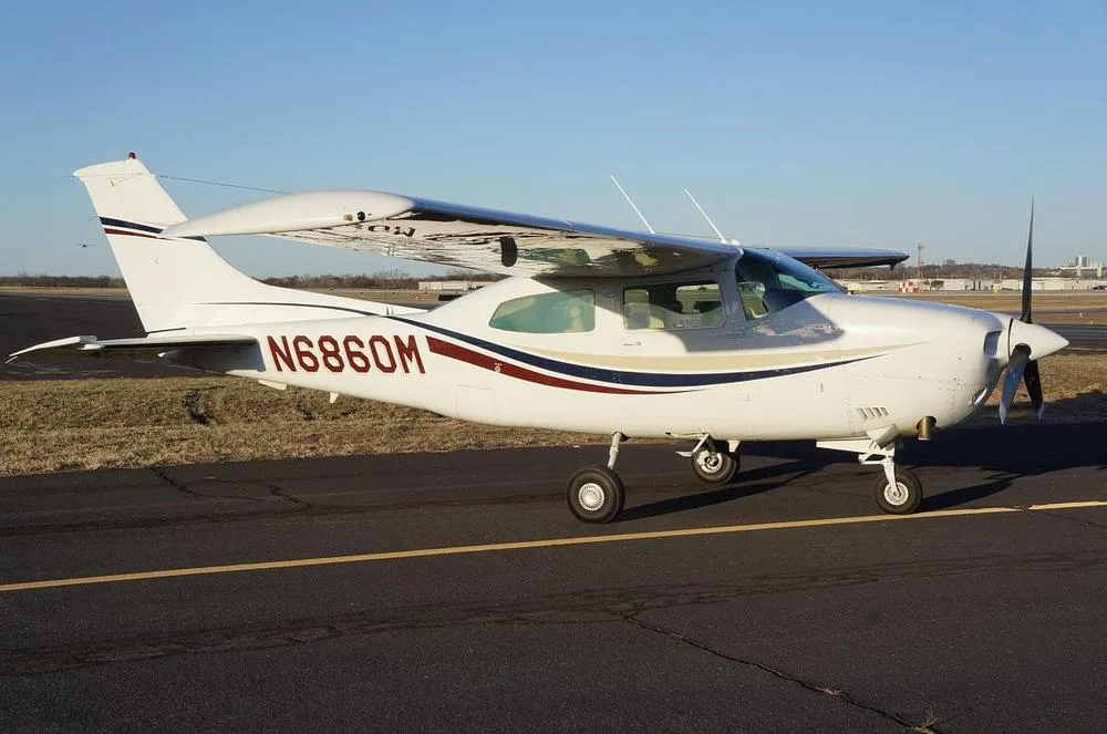 A white Cessna 210 Centurion aircraft with red, blue, and gold stripes on an airfield under a clear blue sky.