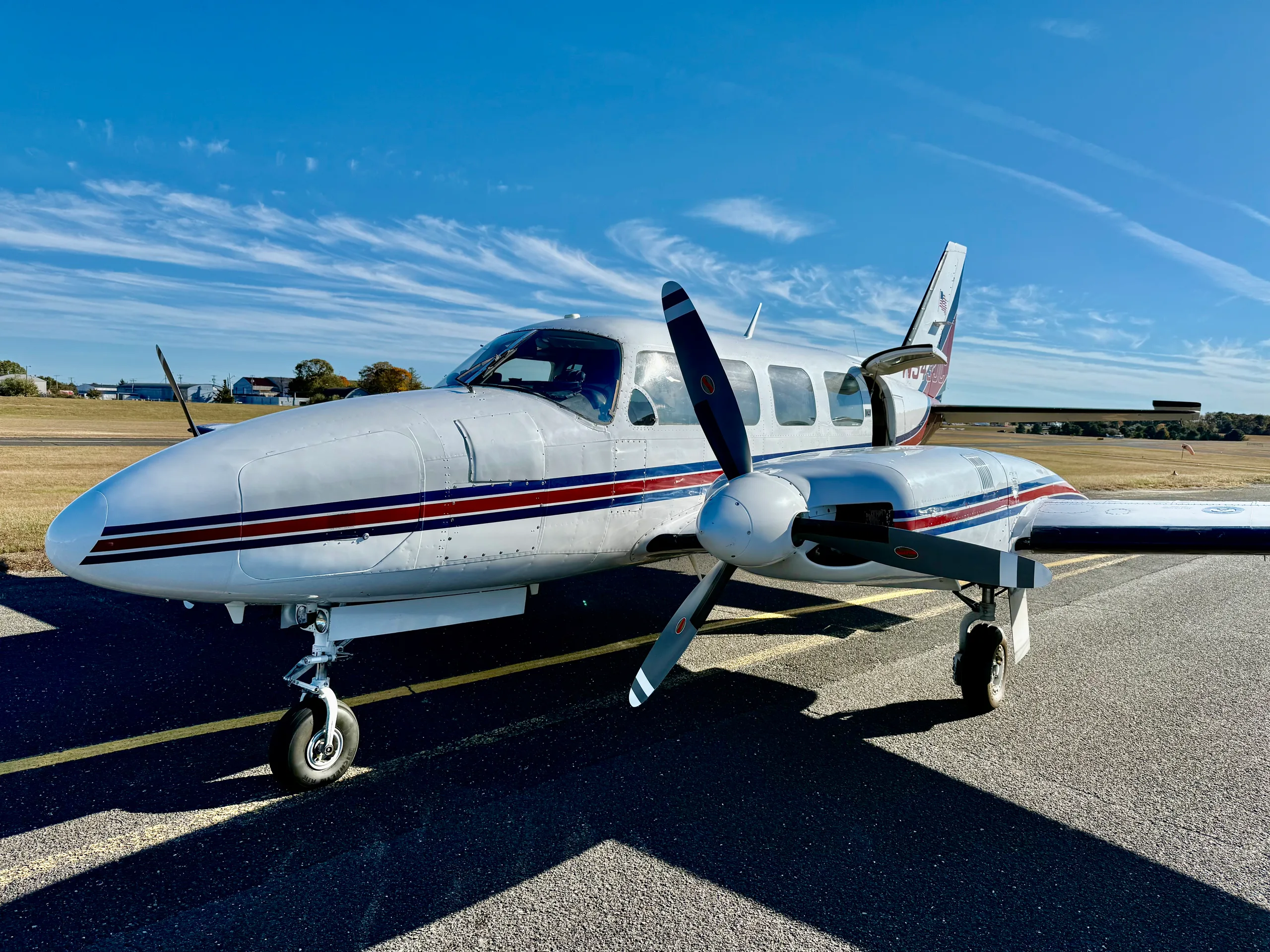 A white twin-engine propeller aircraft with red and blue stripes on the fuselage and tail, parked on an asphalt ramp under a clear blue sky.