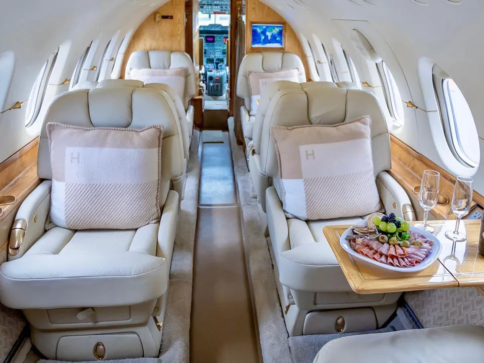 The luxurious interior cabin of a private jet with cream leather seats, wood paneling, and a table set with snacks and drinks.