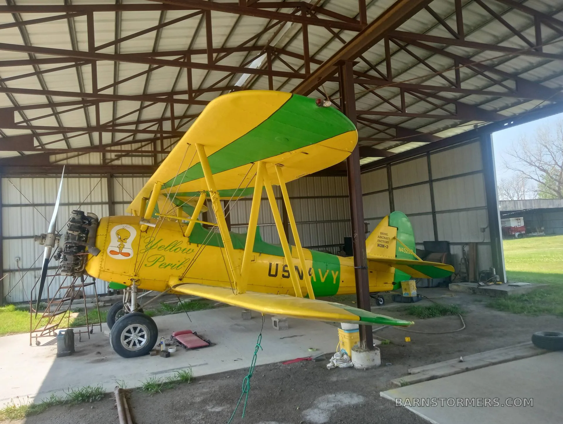 A yellow and green N3N biplane, labeled "US NAVY" and "Yellow Peril" with a Tweety Bird cartoon, is parked in a hangar.