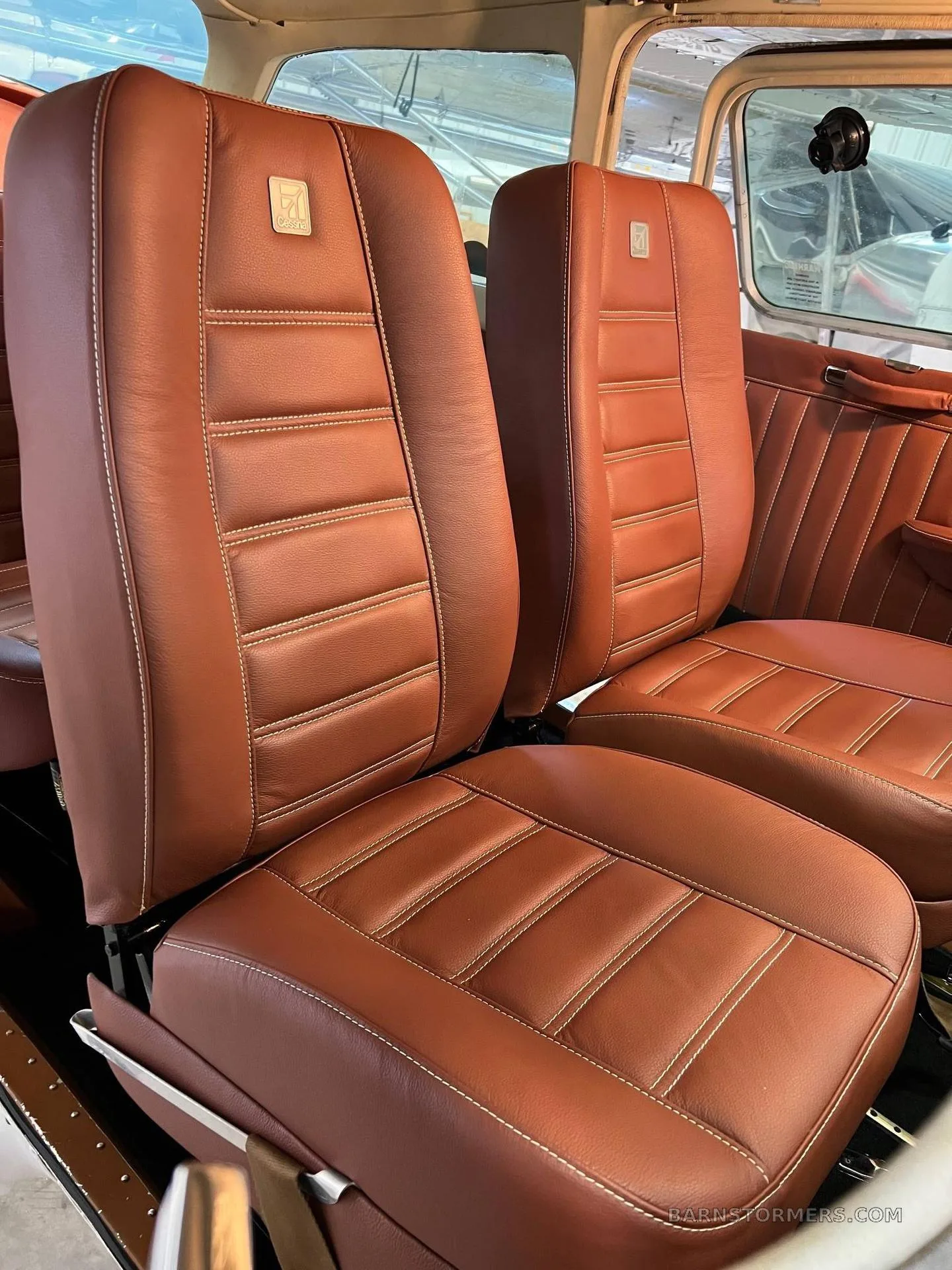 The interior of an aircraft cabin, showing two brown leather seats with white stitching and a small emblem on the headrest.
