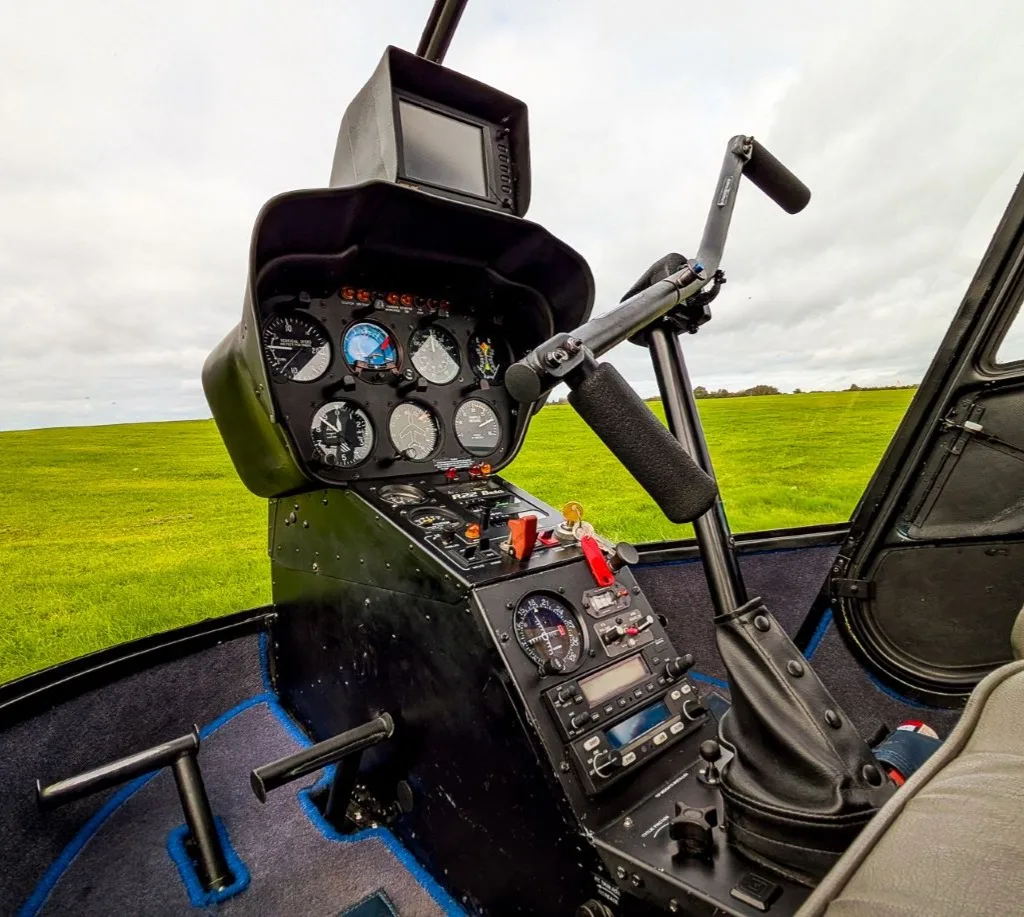 The cockpit of a helicopter showing the instrument panel, cyclic, and collective controls, with a green field outside.