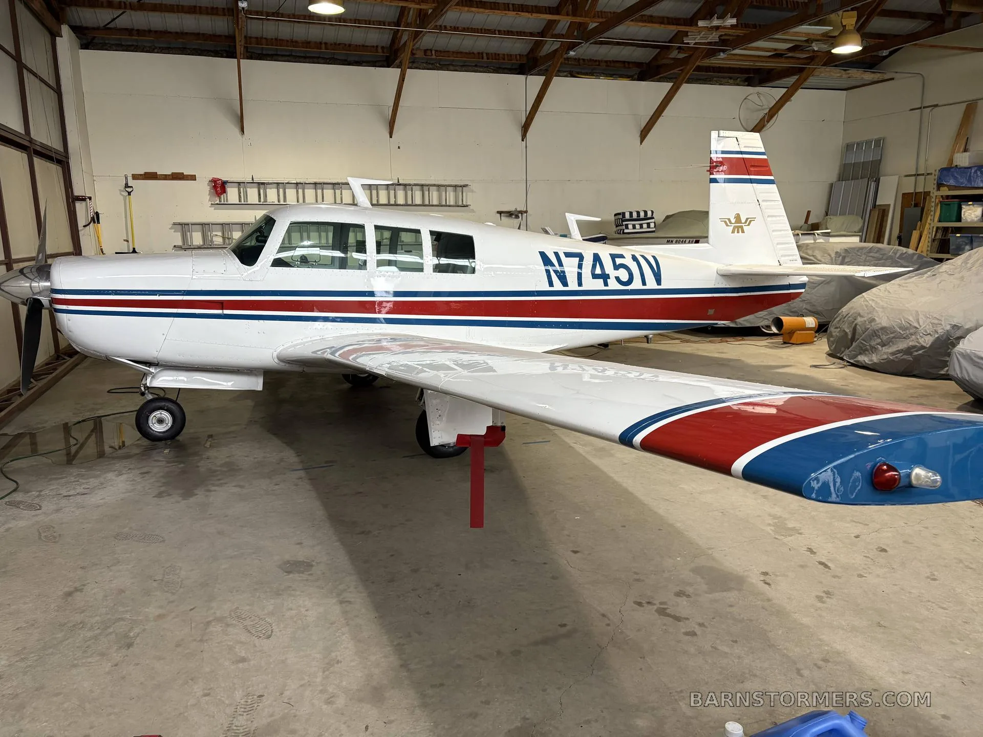 A white single-engine aircraft with red and blue stripes, registration N7451V, parked inside a hangar.