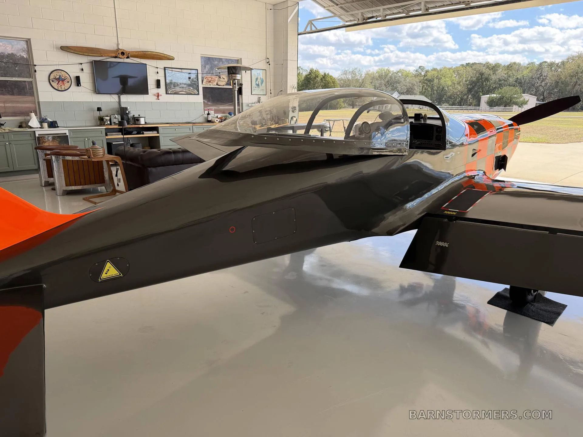 A side view of the black and orange single-engine aircraft with a bubble canopy, parked inside a hangar.