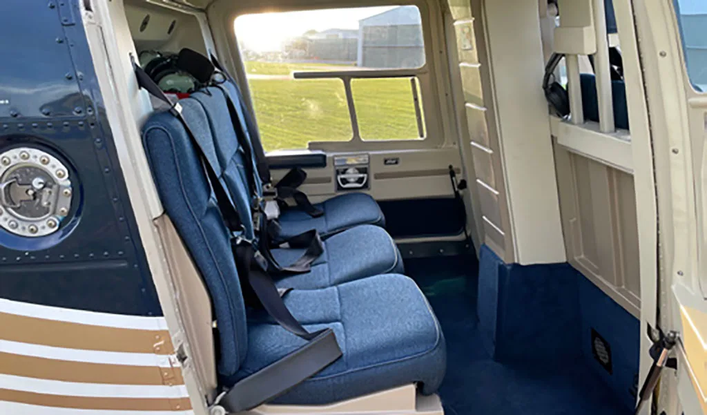 The interior cabin of a helicopter with three blue fabric seats and a view of a grassy field outside.