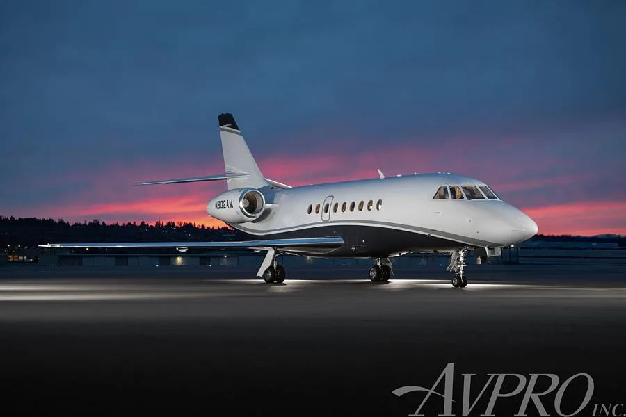 A Dassault Falcon business jet, registration N902AM, parked on an airfield at sunset, showing its white and dark blue paint scheme.