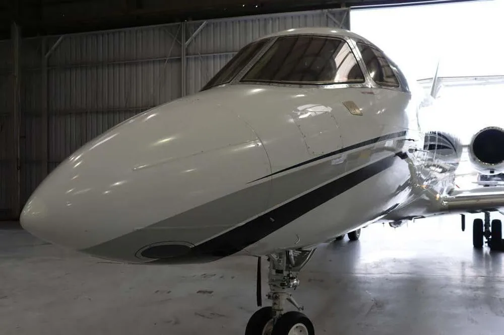 A close-up front view of the white business jet's nose with black and grey stripes, inside a hangar.