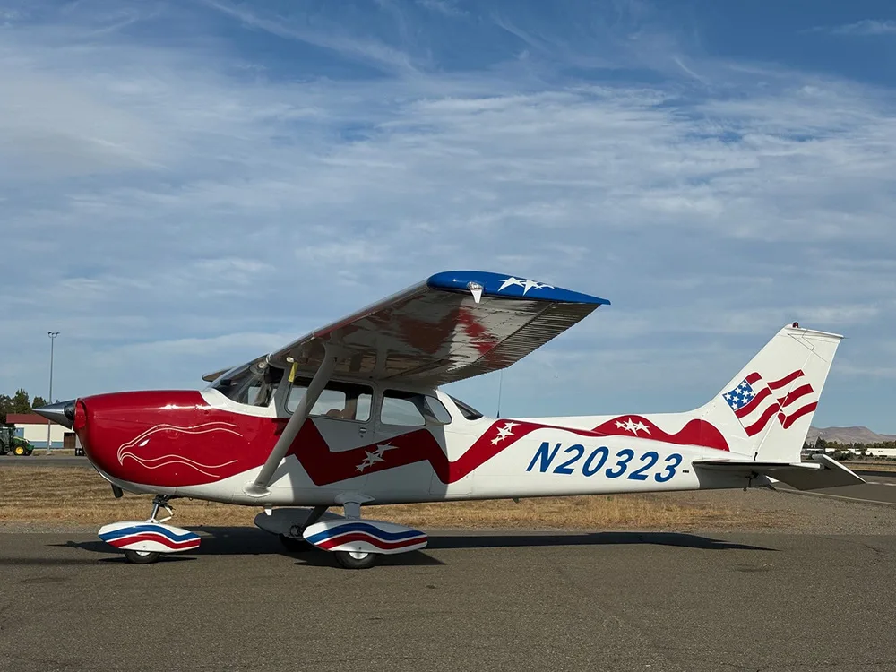 A three-quarter front view of the red, white, and blue Cessna 172, N20323, parked on an airfield under a partly cloudy sky.