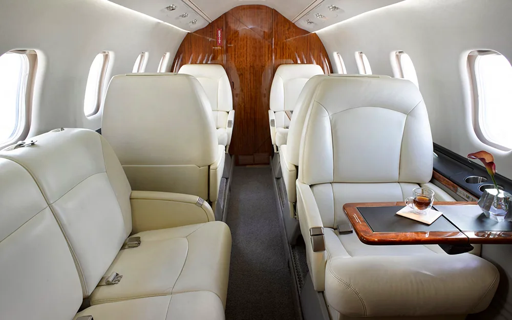 The luxurious interior cabin of a private jet with cream leather seats, a wooden bulkhead, and a table with a drink.