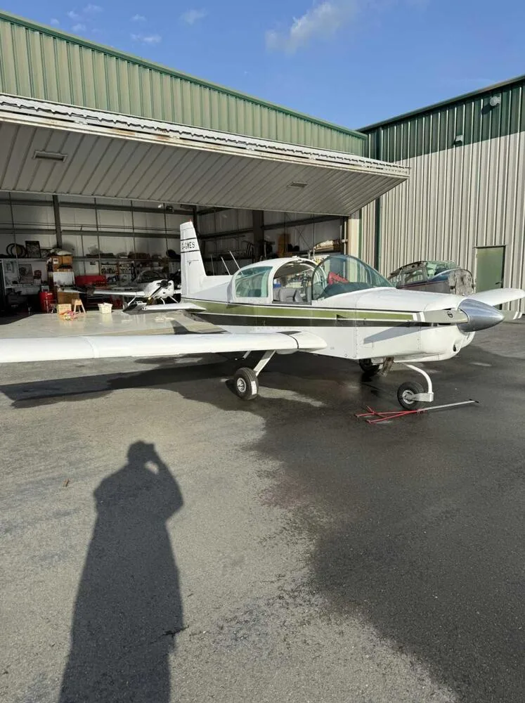 Side view of a white and green Grumman American AA-5 Traveler aircraft with its canopy open, parked outside a hangar.