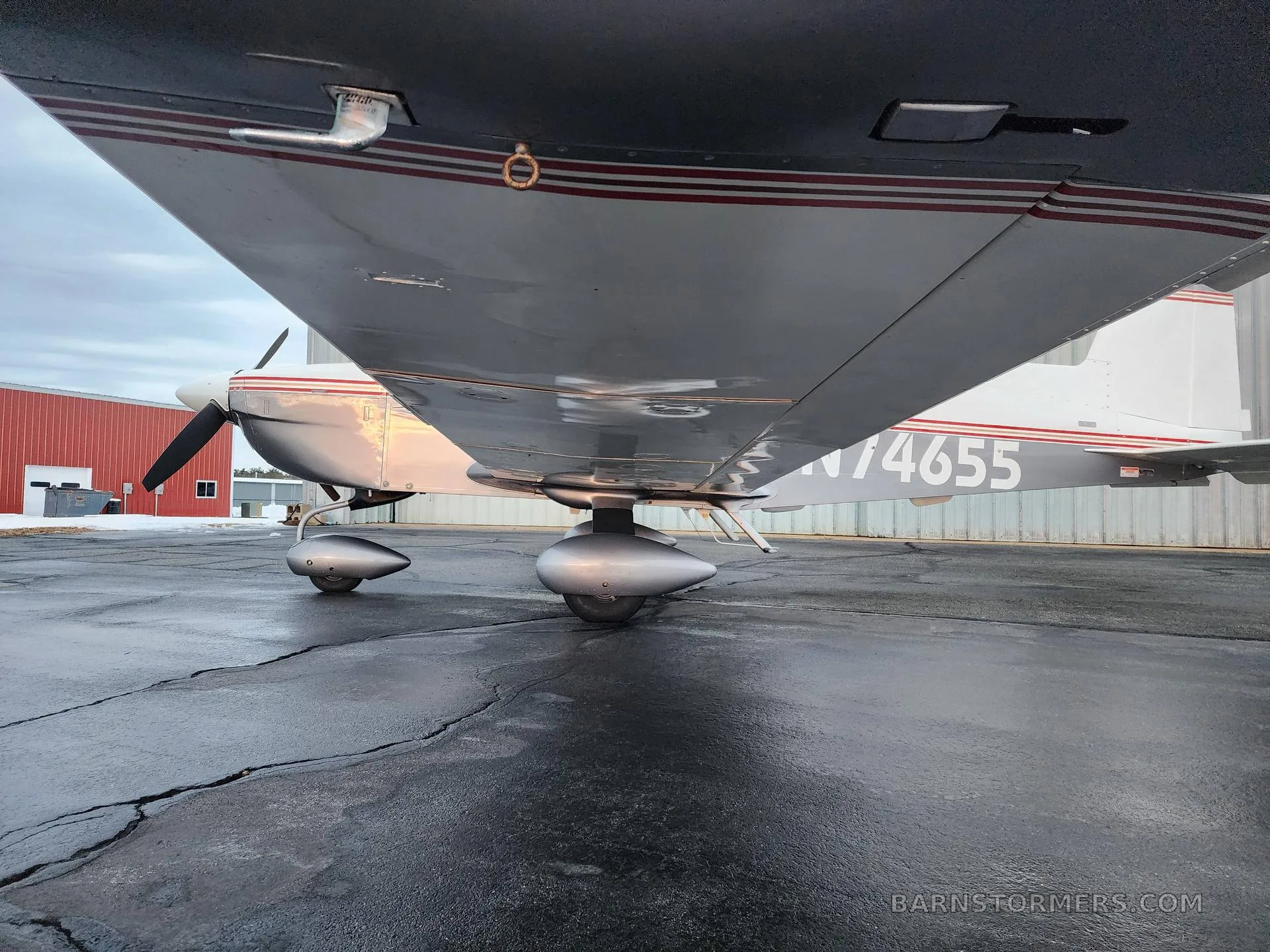 An underside view of the left wing and landing gear of a white and silver Grumman AA-5 Traveler aircraft on a wet tarmac.