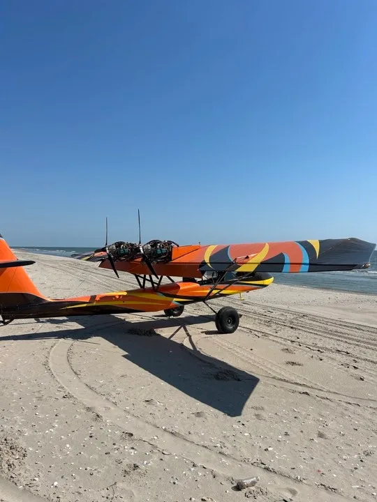 A side view of an orange and black ultralight aircraft with twin engines, parked on a sandy beach next to the ocean.