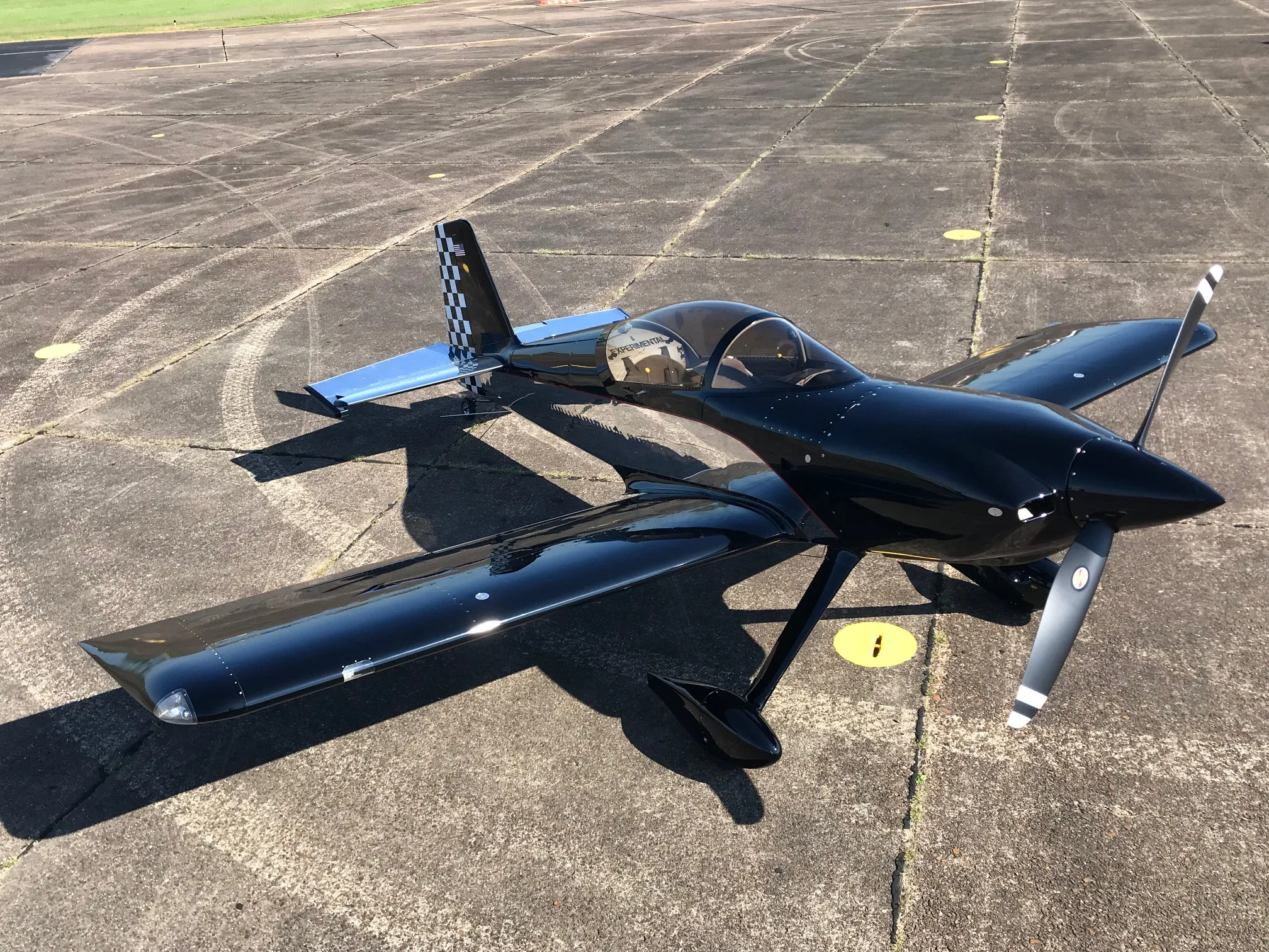 A black single-engine experimental aircraft with a checkered tail and polished silver horizontal stabilizers parked on a tarmac.