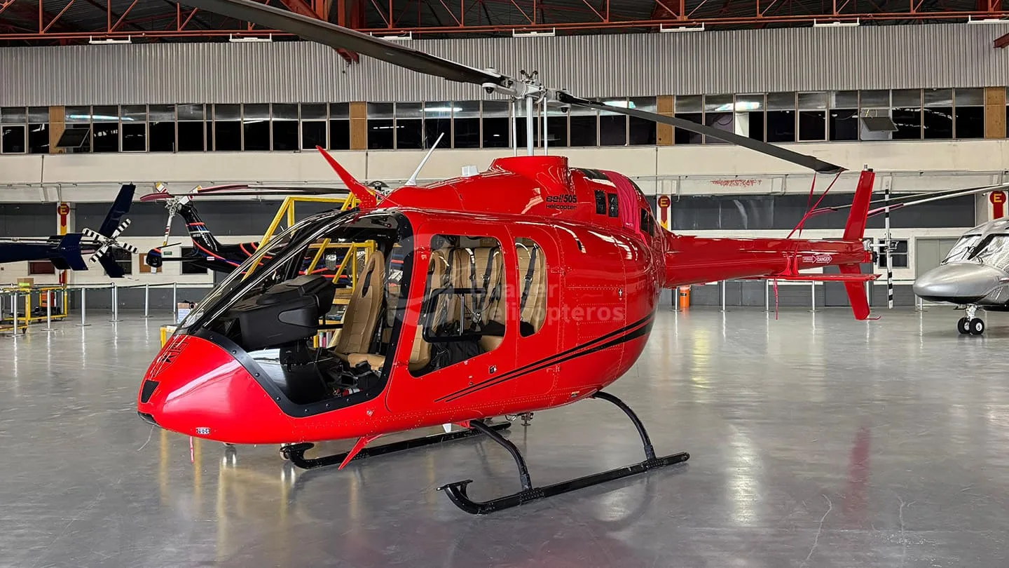 A bright red Bell 505 helicopter with black accents is parked inside a hangar, viewed from the front-side.