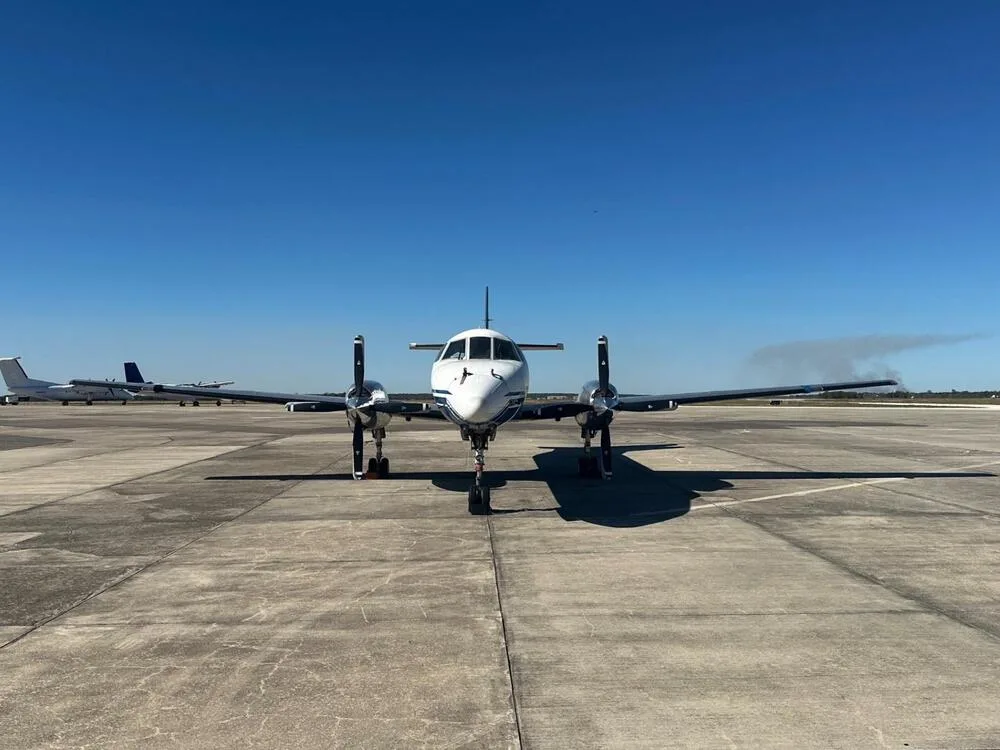 A white and blue Beechcraft 1900 twin-engine turboprop aircraft, registration N-957BA, viewed head-on on an airfield.