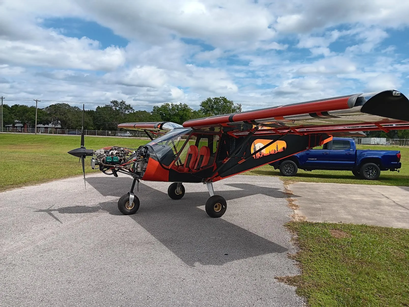 A full view of an orange and black ultralight aircraft with an exposed engine and desert sunset mural, parked on an airfield tarmac.