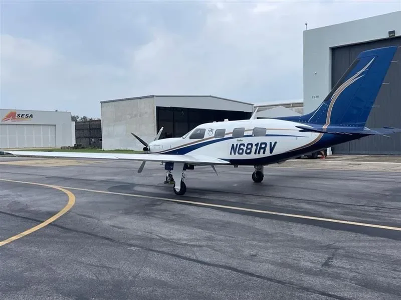 A white and blue twin-engine turboprop aircraft, N681RV, is parked on an airfield with hangars in the background under a cloudy sky.