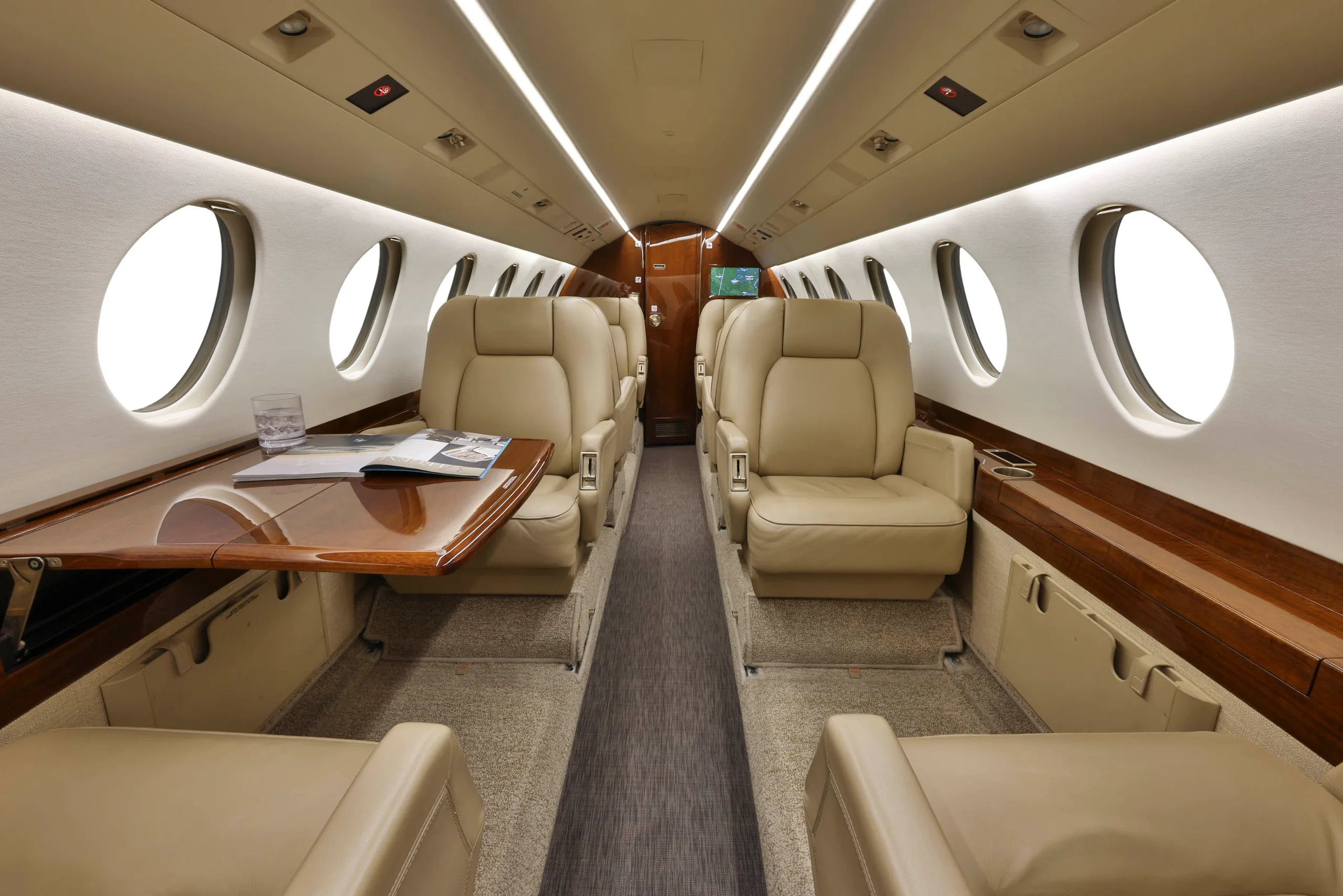 The luxurious interior cabin of a business jet features beige leather seats, a polished wooden table, and large windows.