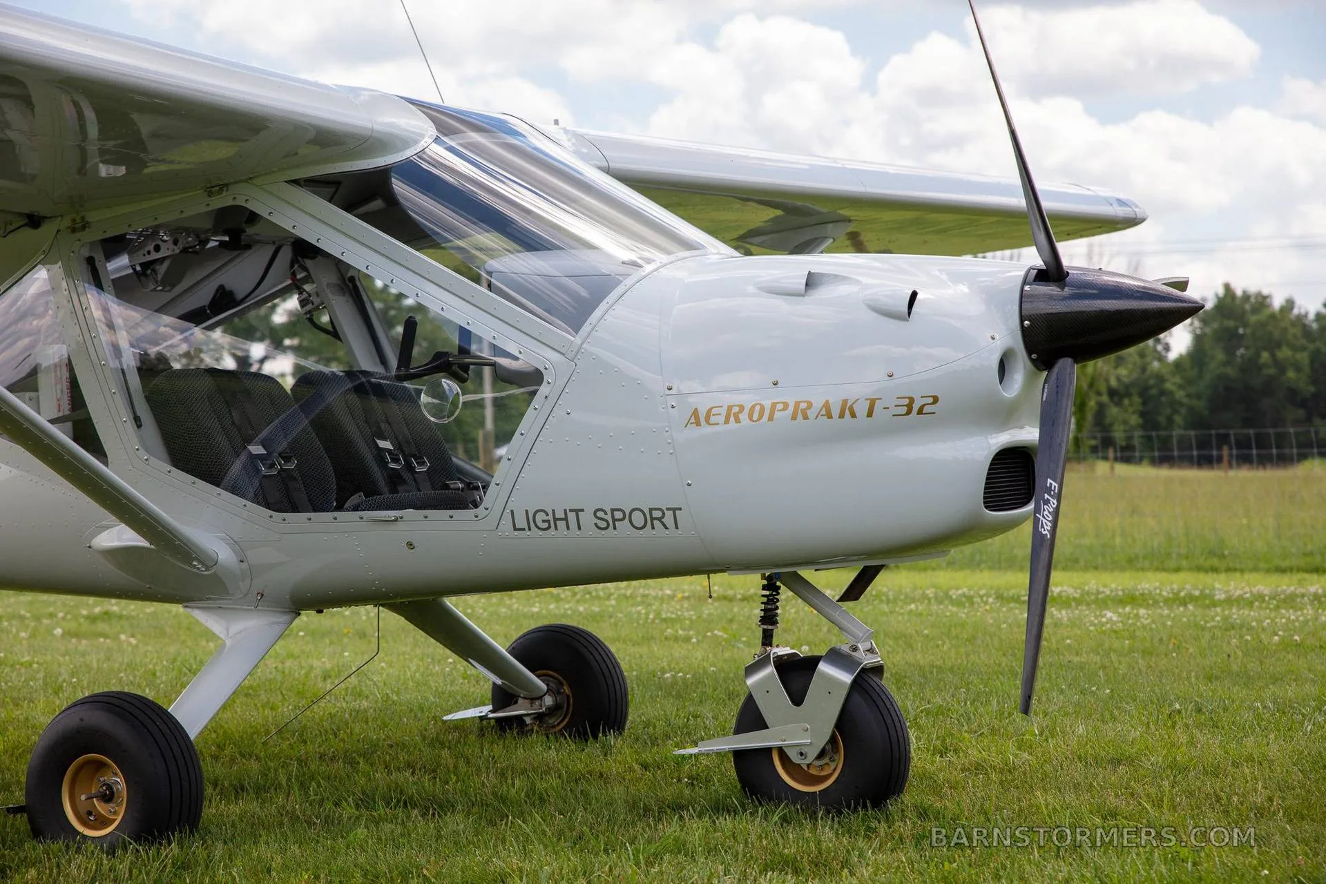 A close-up view of the front right side of a white Aeroprakt-32 light sport aircraft, showing the propeller and cockpit.